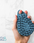 A hand holding a blue, textured scrubber with protruding nubs, identified as The scrub-dub® Scalp and Body Scrubber by Scrub-dub®, perfect for cleaning and exfoliating, against a blurred white background. The logo "scrub-dub®" is in the bottom left corner.