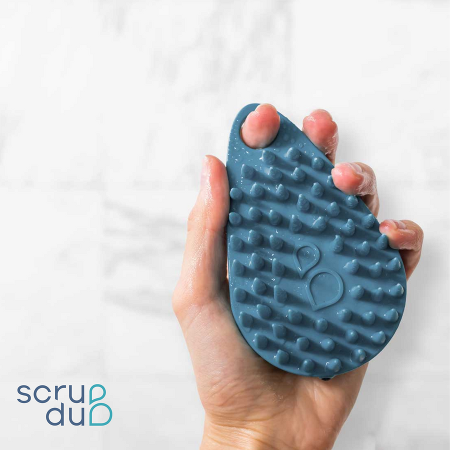 A hand holding a blue, textured Scrub-dub™ Scalp and Body Scrubber with the logo "scrub dub" at the bottom right corner against a white background.