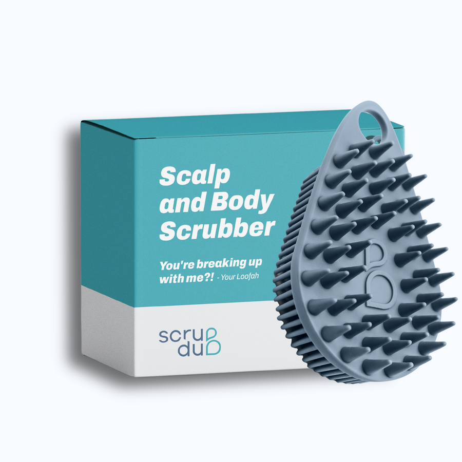 A blue "Scrub-dud™" scalp and body scrubber with gentle bristles, next to its packaging with the text "scalp and body scrubber - you're breaking up with me?!