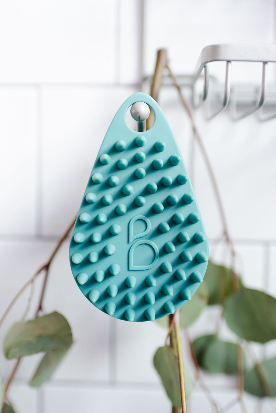 Scrub-dub™ body scrubber hanging from shower hook with eucalyptus in the background.