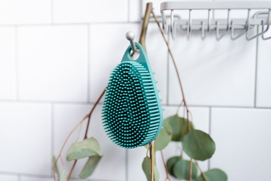 Teal body scrubber handing from shower hook with eucalyptus behind it. 