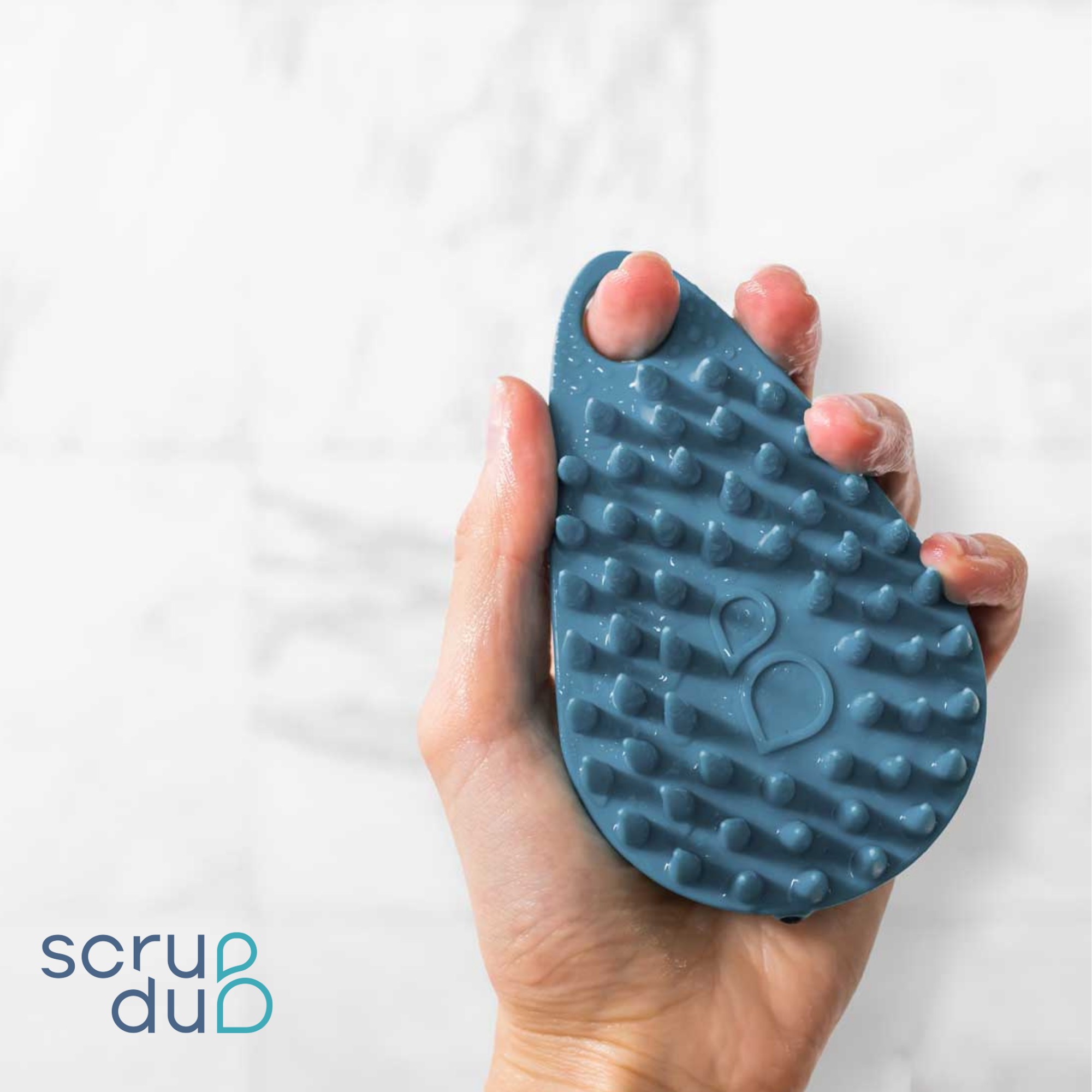 A hand holding a blue, textured scrubber with protruding nubs, identified as The scrub-dub® Scalp and Body Scrubber by Scrub-dub®, perfect for cleaning and exfoliating, against a blurred white background. The logo &quot;scrub-dub®&quot; is in the bottom left corner.