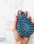 A hand holding a blue, textured Scrub-dub™ Scalp and Body Scrubber with the logo "scrub dub" at the bottom right corner against a white background.