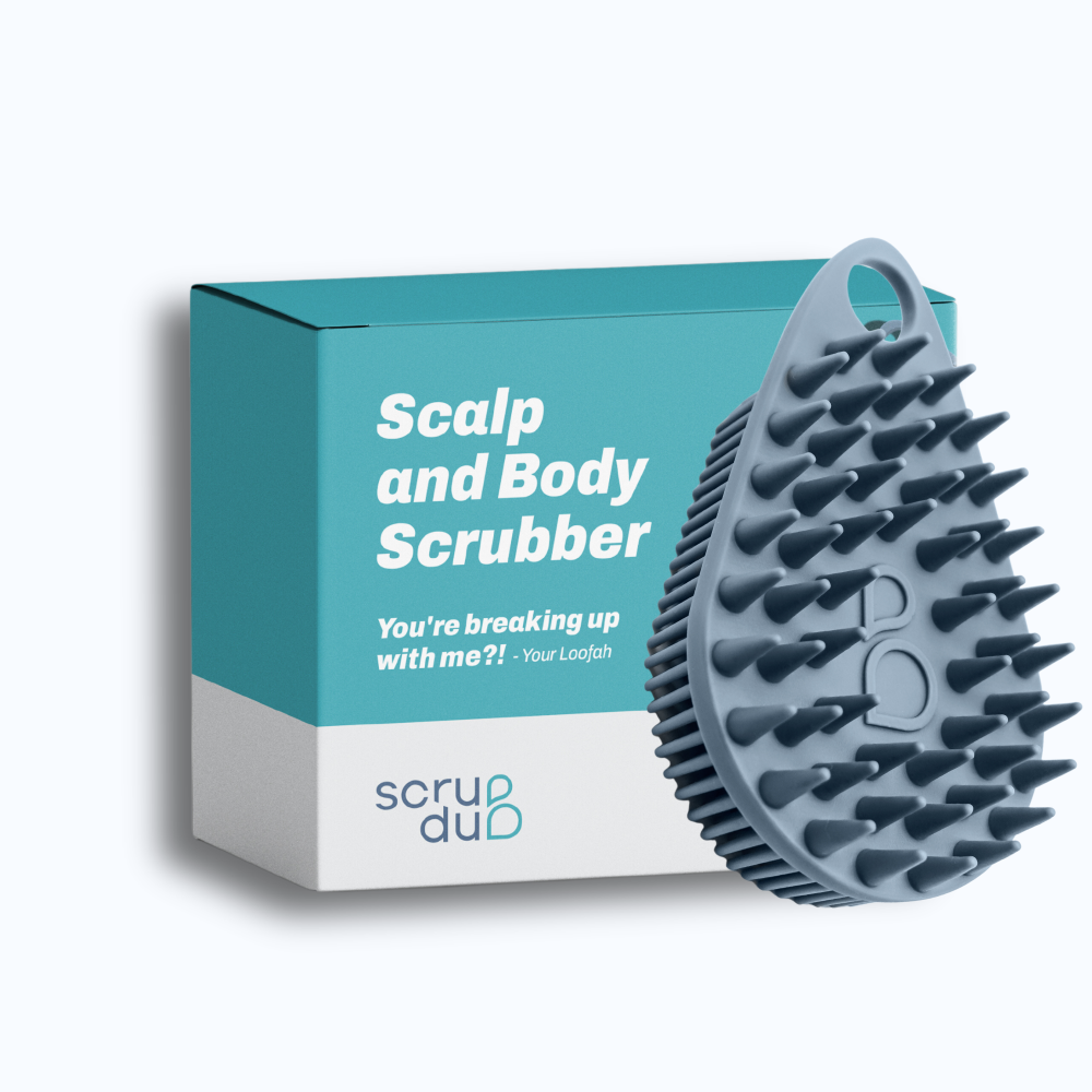 A blue silicone exfoliating brush is displayed in front of its teal and white packaging, which reads &quot;The scrub-dub® Scalp and Body Scrubber&quot; and &quot;You&#39;re breaking up with me?! – Your Loofah.&quot; The brand name, Scrub-dub®, is located at the bottom.