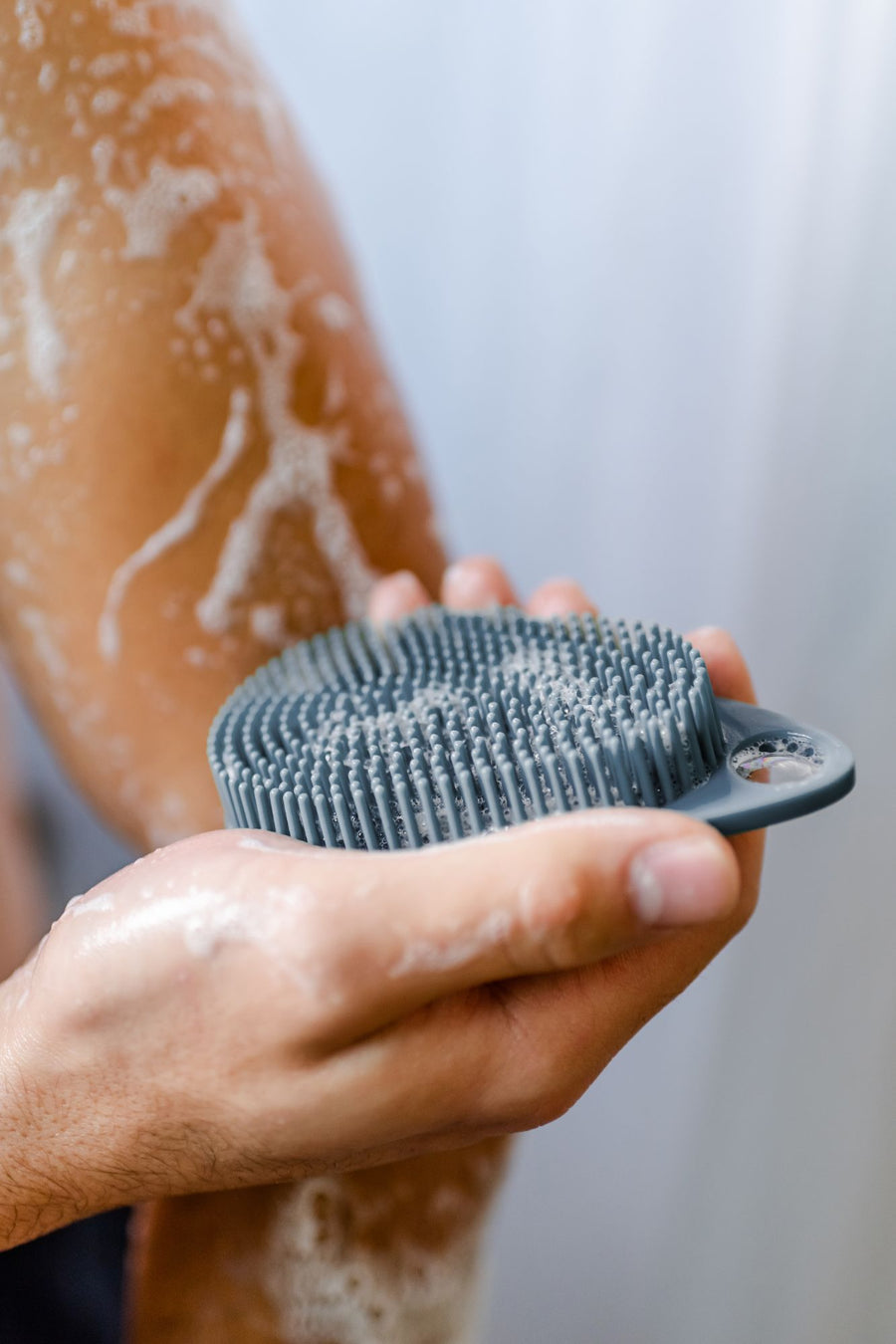 Man in shower holding silicone body scrub with soap on scrub and aarm.