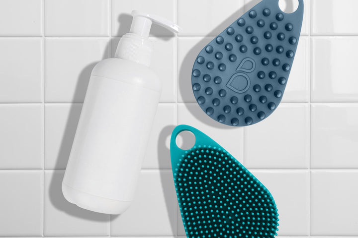 Two silicone body scrubs with a white bottle of soap against a white tile background.